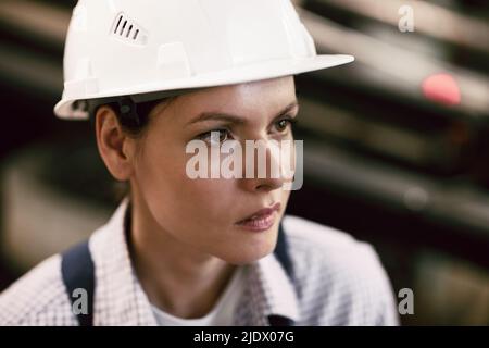 Nahaufnahme einer ernsthaften, brutalen jungen Frau in Hardhat, die in der Fabrik arbeitet und industrielle Prozesse in Erwägung zieht Stockfoto