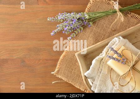 Natürliches Lavendel-Seifenstück auf Handtuch in Holzschale auf Holztisch mit einem Strauß Lavendelblüten-Stacheln. Draufsicht. Horizontale Zusammensetzung. Stockfoto