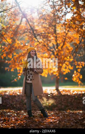 Hallo oktober. Ganzkörperportrait einer glücklichen, stilvollen Frau mittleren Alters in braunem Mantel und gelbem Hut mit herbstlichen gelben Blättern draußen im Stadtpark Stockfoto