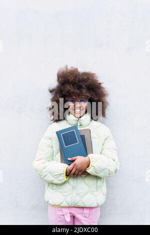 Schulmädchen hält Bücher, die vor einer weißen Wand stehen Stockfoto