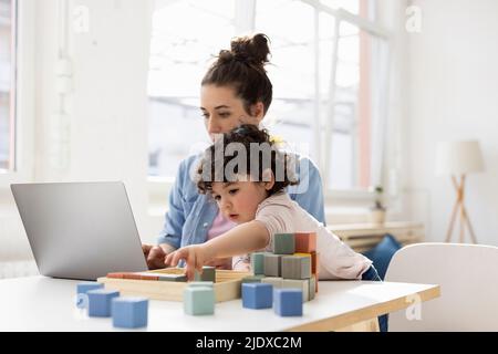 Mutter, die von zu Hause aus mit einem Laptop arbeitet, während ihre Tochter mit Bausteinen spielt Stockfoto