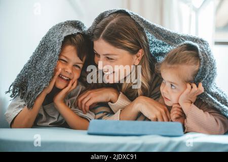 Lächelnde junge Frau und Mädchen, die den Jungen unter der Decke auf dem Bett liegen sehen Stockfoto