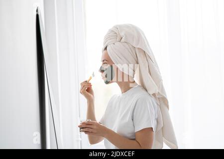 Junge Frau, die zu Hause Gesichtsmaske vor dem Spiegel aufführt Stockfoto