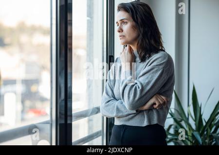 Nachdenkliche traurige Frau, die zu Hause durch das Fenster schaut Stockfoto