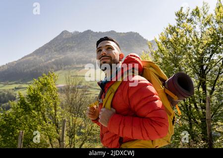 Lächelnder Wanderer im Rucksack, der an sonnigen Tagen vor dem Baum steht Stockfoto