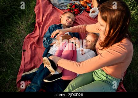 Mutter kitzelt Kinder liegen auf Picknickdecke auf dem Feld Stockfoto