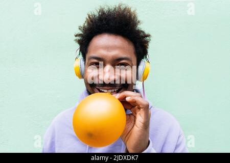 Lächelnder Mann hört Musik über Kopfhörer, der vor türkisfarbener Wand einen Ballon bläst Stockfoto