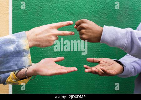 Hände von Freunden, die vor einer grünen Wand eine Schere aus Steinpapier spielen Stockfoto