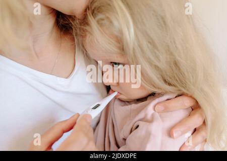 Mutter trägt Tochter und misst Temperatur mit Thermometer zu Hause Stockfoto