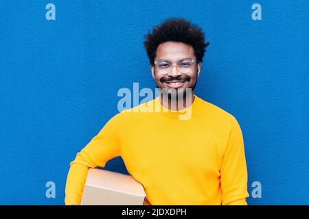 Lächelnder Delivery-Mann hält Box vor der blauen Wand Stockfoto