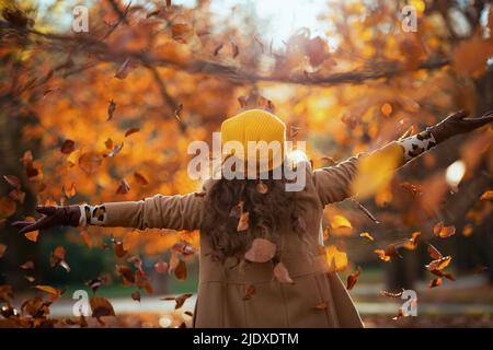 Hallo september. Von hinten gesehen Frau in beigem Mantel und orangefarbenem Hut, die im Herbst im Stadtpark jubelt. Stockfoto