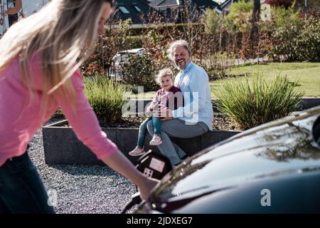 Lächelnder Mann mit Tochter, die Frau beim Laden des Autos an einem sonnigen Tag ansah Stockfoto