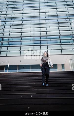 Lächelnde Geschäftsfrau, die auf der Treppe vor dem modernen Gebäude nach unten geht Stockfoto