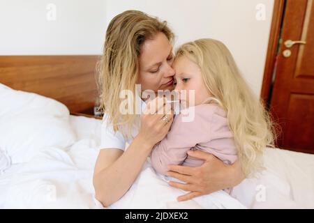 Mutter trägt Tochter und misst Temperatur mit Thermometer im Schlafzimmer Stockfoto