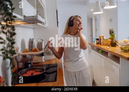 Schöne junge Frau mit Kopfhörern, die in der Küche Musik hört. Pfannkuchen braten. Stockfoto