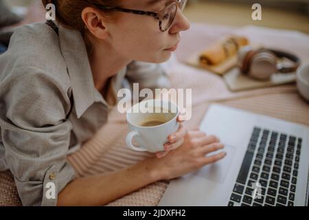 Frau trägt Brillen und hält Kaffee Tasse E-Learning über Laptop zu Hause Stockfoto