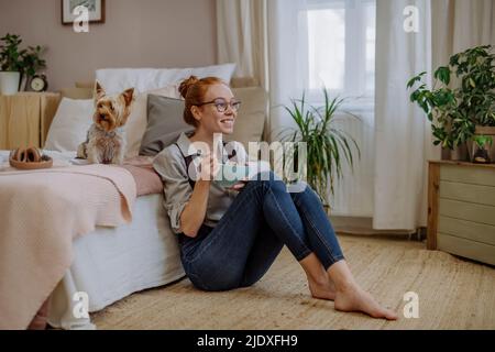 Glückliche Frau mit einer Schüssel mit Essen, die zu Hause beim Hund auf dem Bett sitzt Stockfoto