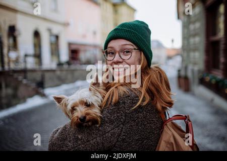 Glückliche Rothaarige Frau trägt Strickmütze trägt Yorkshire Terrier in der Stadt Stockfoto