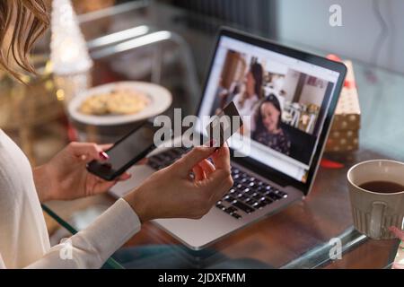 Frau mit Kreditkarte und Smartphone macht Videoanruf mit Freunden über Laptop zu Hause Stockfoto