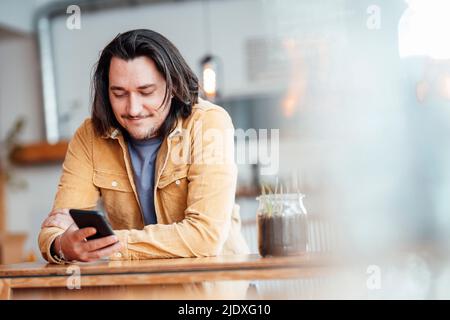 Lächelnder Mann mit langen Haaren, der sich auf den Tisch lehnte und im Café ein Smartphone benutzte Stockfoto