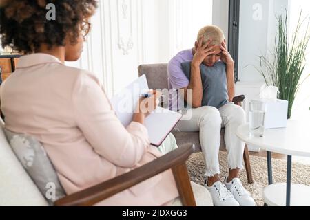 Gestresster Mann, der am Arbeitsplatz mit dem Kopf in den Händen des Psychologen im Sessel sitzt Stockfoto