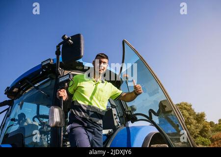 Junger Bauer, der den Daumen hoch stund an der Tür des Traktors Stockfoto