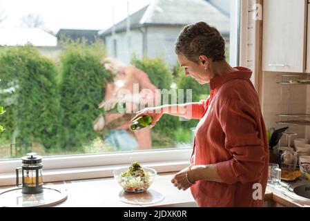 Frau am Fenster in der Küche gießt Olivenöl in die Salatschüssel Stockfoto