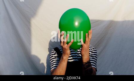 Mann bedeckt Gesicht mit grünen Ballon vor weißem Hintergrund Stockfoto