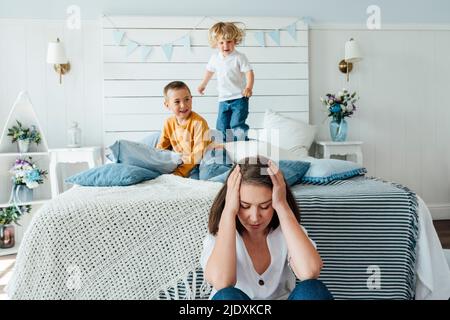 Jungen spielen auf dem Bett von müde Frau sitzen mit geschlossenen Augen zu Hause Stockfoto