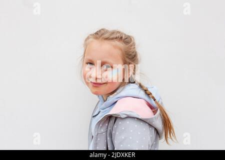 Lächelndes Mädchen mit ukrainischer Flagge auf Wange vor weißem Hintergrund gemalt Stockfoto