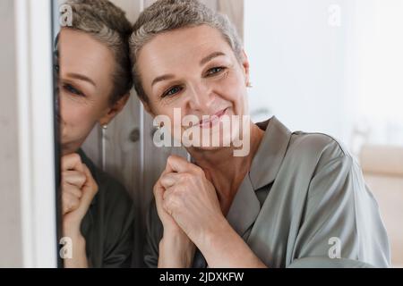 Porträt einer reifen Frau mit kurzen grauen Haaren, die sich gegen den Spiegel lehnt Stockfoto