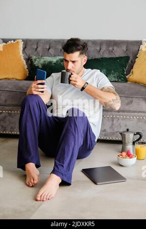 Ein junger Mann mit dem Mobiltelefon trinkt Kaffee und sitzt zu Hause vor dem Sofa Stockfoto