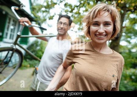 Glückliche reife Frau mit Mann, der Fahrrad auf der Schulter im Hinterhof trägt Stockfoto