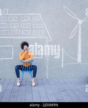 Nachdenkliche Frau, die auf einem Stuhl vor der Wand mit bemalter Windturbine und Haus sitzt Stockfoto