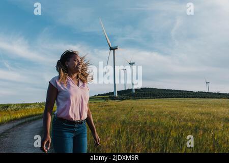 Lächelnde Frau mit zerzausten Haaren, die vor der Windturbine steht Stockfoto