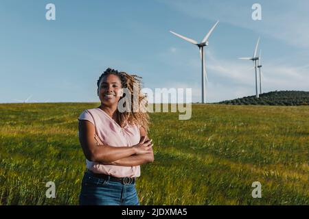 Lächelnde junge Frau, die an sonnigen Tagen mit gekreuzten Armen auf der Wiese steht Stockfoto