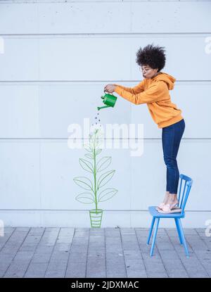 Frau, die auf einem Stuhl steht und Blumen an der Wand bewässert Stockfoto