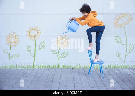 Frau, die auf einem Stuhl steht und Blumen an der Wand bewässert Stockfoto