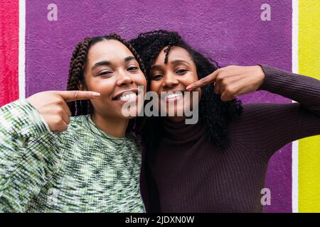 Glückliche Freunde zeigen sich gegenseitig mit den Fingern, die vor einer mehrfarbigen Wand stehen Stockfoto