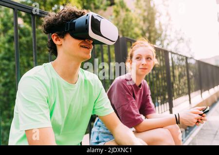 Lächelnder Junge im Virtual-Reality-Simulator, der von einem Freund vor dem Geländer sitzt Stockfoto