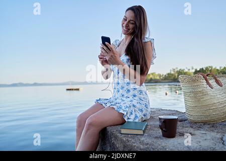 Glückliche Frau, die auf dem Steg am See mit dem Handy telefonierte Stockfoto