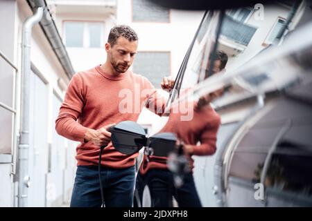 Mann, der in der Nähe des Gebäudes ein Elektroauto mit Kabel aufgeladen hat Stockfoto