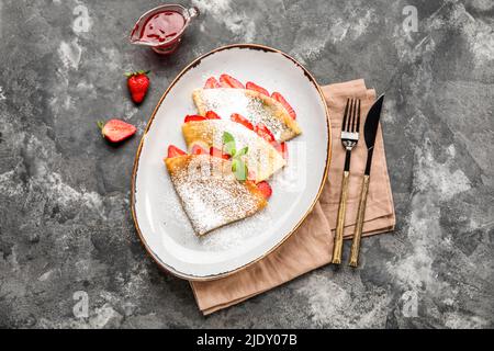 Teller mit dünnen Pfannkuchen mit Erdbeeren, Serviette und Besteck auf dunklem Hintergrund Stockfoto
