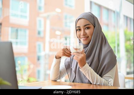 Attraktive und fröhliche asiatische muslimische Arbeiterin oder College-Studentin mit Hijab-Fernbedienung, die im Café mit einer Kaffeetasse arbeitet. Stockfoto