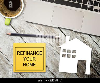 Text mit Refinanzierung Your Home Schreiben Sie auf eine gelbe Haftnotiz und Heimpapier, Laptop-Computer, Bleistift, Tasse Kaffee auf Holztisch Hintergrund. Hypothek c Stockfoto