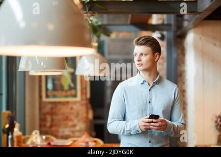 Seriös nachdenkliche junge Managerin im lässigen Hemd, die im modernen Café steht und nachdenklich davonschaut, während sie SMS am Telefon schreibt Stockfoto
