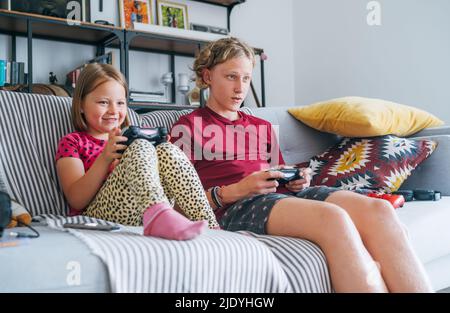 Lächelnde kleine Schwester mit Bruder sitzt auf dem Sofa im Wohnzimmer, schaut Fernsehen und spielt Konsole Videospiel mit Wireless-Controller. Nachlässiger Chil Stockfoto