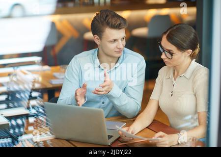 Seriöse junge Unternehmer, die im modernen Café am Tisch sitzen und mit einem Laptop über Daten diskutieren Stockfoto