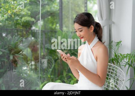 Das asiatische Mädchen sitzt am Tisch neben dem Fenster, hält ein Mobiltelefon und schaut auf den Bildschirm Stockfoto