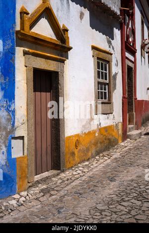 Dekorativ bemalte Giebel schmücken Granittüren in einer ruhigen gepflasterten Straße mit traditionellen portugiesischen Häusern in der historischen mittelalterlichen ummauerten Stadt Óbidos, Centro, Portugal. Stockfoto